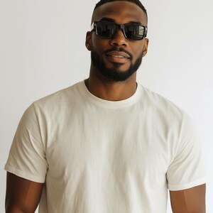 K&ouml;nnte beinhalten: Eine Person tr&auml;gt eine schwarze Sonnenbrille und ein cremefarbenes T-Shirt. Die Sonnenbrille hat eine rechteckige Form mit dicken Rahmen. Das T-Shirt ist schlicht und hat kurze &Auml;rmel. Der Hintergrund ist einfarbig wei&szlig;.