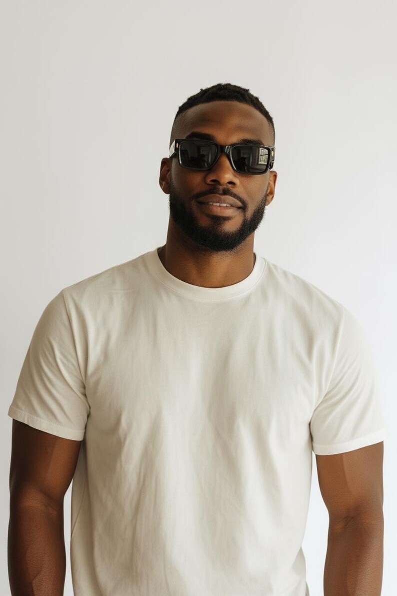 K&ouml;nnte beinhalten: Eine Person tr&auml;gt eine schwarze Sonnenbrille und ein cremefarbenes T-Shirt. Die Sonnenbrille hat eine rechteckige Form mit dicken Rahmen. Das T-Shirt ist schlicht und hat kurze &Auml;rmel. Der Hintergrund ist einfarbig wei&szlig;.