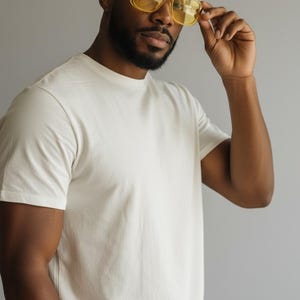 May include: A person wearing a cream-colored, short-sleeved t-shirt and yellow-tinted square sunglasses. The person is holding the sunglasses with their right hand. The background is a solid gray color.
