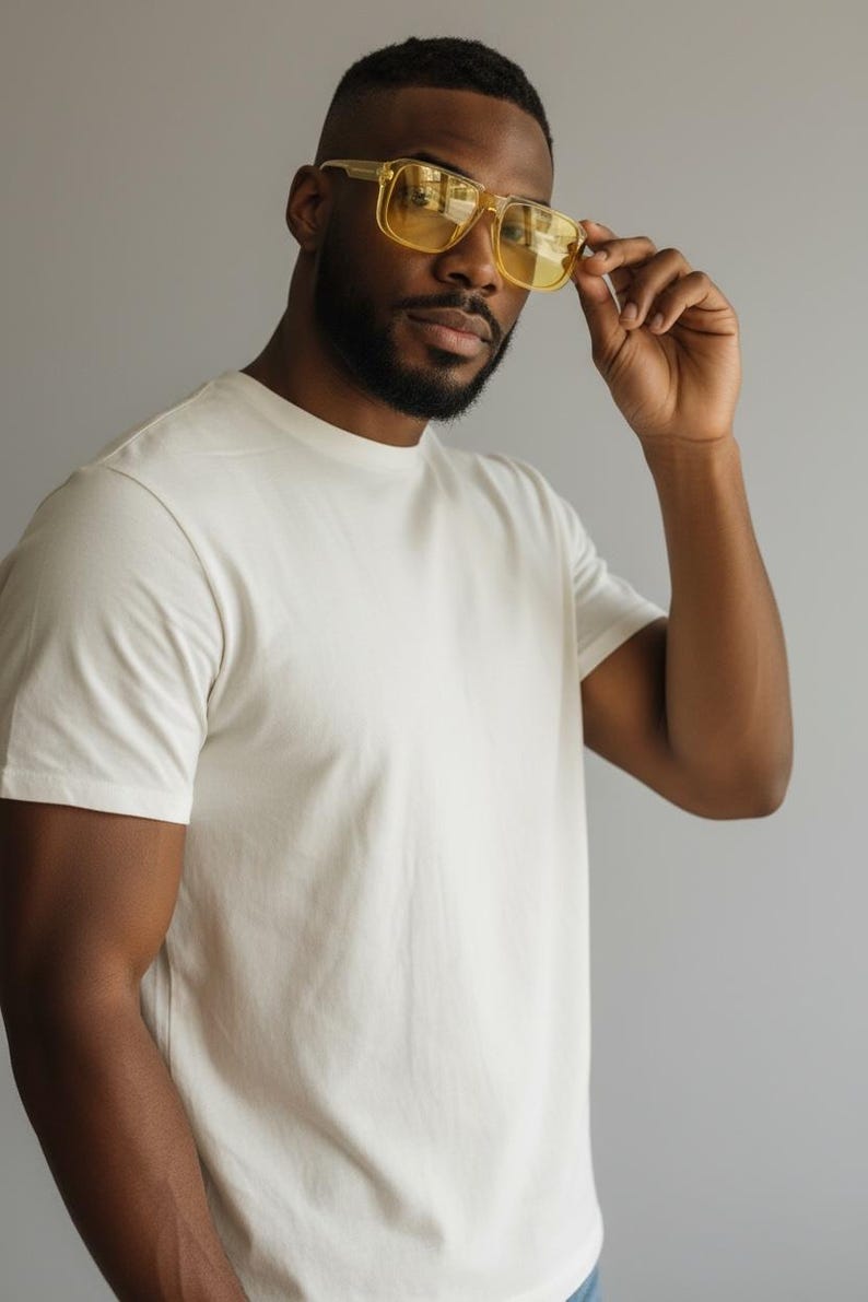 May include: A person wearing a cream-colored, short-sleeved t-shirt and yellow-tinted square sunglasses. The person is holding the sunglasses with their right hand. The background is a solid gray color.