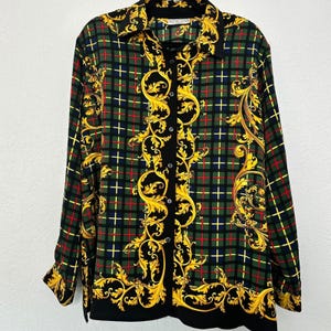May include: A long-sleeved button-down shirt with a black, green, and red plaid pattern. The shirt features gold baroque-style scrollwork and a black collar and cuffs. The shirt has a classic collar and button-up front.