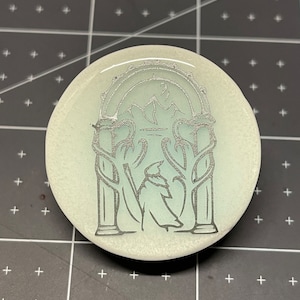 Könnte beinhalten: Ein runder Untersetzer aus weißer Keramik mit einem silbernen Design eines Torbogens mit einer stehenden Figur in der Mitte. Der Torbogen hat zwei Säulen und eine gewölbte Oberseite. Die Figur trägt eine lange Robe und einen spitzen Hut.