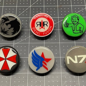May include: Six round, white, plastic buttons with different colored and graphic designs. The buttons feature a wolf, a rocket ship, a person with a thumbs up, an umbrella, a winged star, and a logo with the letters N7.