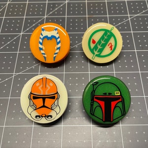 May include: Four round, colorful, pin-back buttons featuring Star Wars characters. The buttons are white, orange, green, and cream with images of a clone trooper, a bounty hunter, a Jedi, and a symbol.