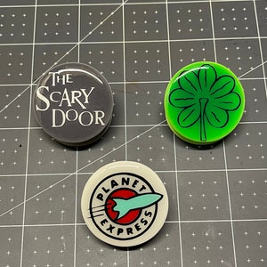 May include: Three round buttons with different designs. The first button is gray with white text that reads "THE SCARY DOOR". The second button is green with a black outline of a four-leaf clover. The third button is white with black text that reads "PLANET EXPRESS" and a red circle with a blue rocket ship.