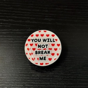 May include: A circular pin featuring a white background covered in small red hearts. The center of the pin displays the text "YOU WILL NOT BREAK ME" in bold, black, capital letters with a white outline, arranged in four stacked lines.