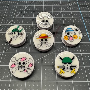 May include: Six white round plugs with different cartoon characters on them. The characters are all from the anime One Piece. The characters are Luffy, Zoro, Sanji, Chopper, Brook, and Franky.