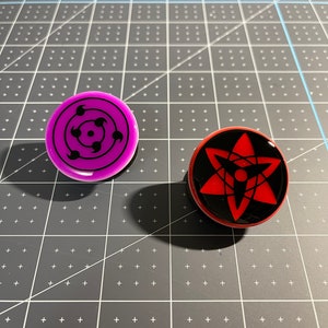 May include: Two round, flat, decorative pins with black backgrounds and colorful designs. The left pin has a purple background with a black and white design of a circle with three concentric circles inside. The right pin has a red background with a black and white design of a star with a circle in the center.