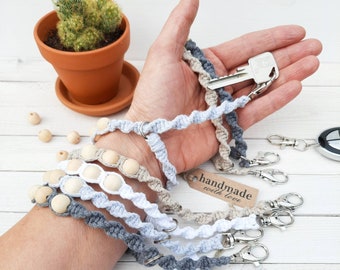 May include: A set of five keychains made with macrame and wooden beads. The keychains are in shades of grey, white, and beige. The keychains are handmade and have a tag that says "handmade with love".