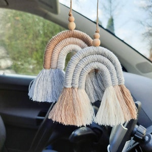 Può includere: Due ciondoli per auto a forma di arcobaleno in macramè con perline di legno. Gli arcobaleni sono realizzati con filato beige, marrone chiaro e grigio. I ciondoli sono appesi allo specchietto retrovisore di un'auto.