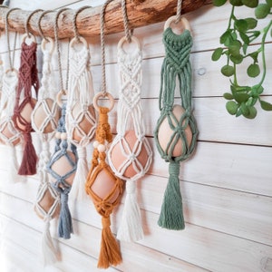 Op de afbeelding: Een verzameling macramé eierrekken met verschillende gekleurde kwastjes. De eierrekken zijn gemaakt van natuurlijke katoenen koord en hebben verschillende patronen.