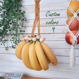 May include: A macrame banana hanger with wooden beads, hanging from a white wooden wall. The hanger is made with a mustard yellow cord and is holding a bunch of ripe bananas. The text "Custom colors" is written on the wall.