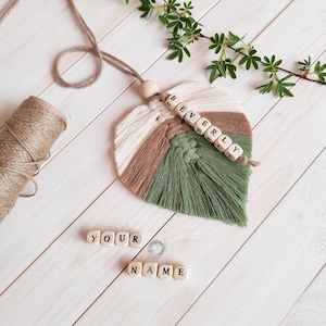 Può includere: Una foglia di macramè verde, marrone e bianca con il nome "BEVERLY" scritto con perline di lettere in legno. La foglia è appesa a un cordoncino marrone. Ci sono anche perline di lettere in legno che compongono "YOUR NAME" su una superficie di legno bianco.
