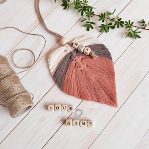 Może przedstawiać: Liść z makramu w kolorze różowym, brązowym i białym z drewnianym koralikiem i imieniem "MIA" napisanym drewnianymi literami. Litery "YOUR NAME" są napisane drewnianymi literami pod liściem.