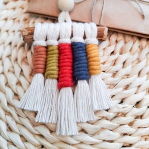 May include: A handmade macrame wall hanging featuring a wooden dowel and colorful tassels. The tassels are in shades of white, orange, yellow, red, and blue. A wooden bead and a tag with text are also visible. The item is displayed on a woven surface.
