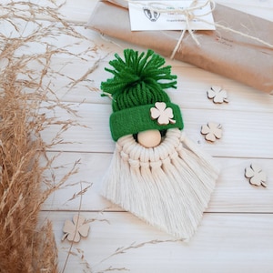 Könnte beinhalten: Ein grüner Garn-Zwerg mit einem weißen Garn-Bart und einem grünen Filzhut mit einem Kleeblatt auf der Vorderseite. Der Zwerg sitzt auf einer weißen Holzoberfläche.