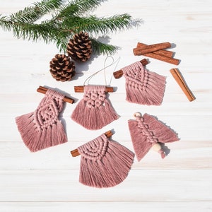 Könnte beinhalten: Fünf rosafarbene Makramee-Anhänger mit Fransen, die jeweils an einem Zimtstange hängen. Die Anhänger haben die Form eines Baumes, eines Kreises und zweier Quadrate.