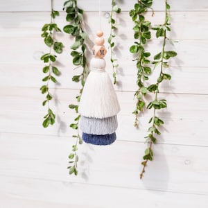 May include: A white, grey, and blue tassel with wooden beads and the letter "M" on each bead. The tassel is hanging from a white wooden wall with green vines.