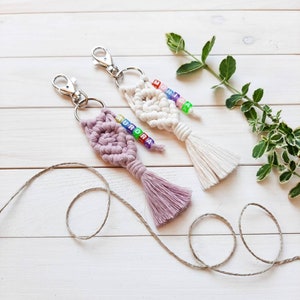 Op de afbeelding: Twee macramé sleutelhangers met kleurrijke kralen met de namen "Aurora" en "Monica", hangend aan een witte houten achtergrond. De sleutelhangers hebben roze en witte kwastjes.