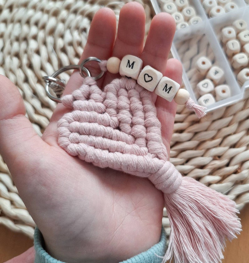 May include: A pink macrame heart keyring with a wooden bead charm that spells "MOM" with a heart bead in between the letters.