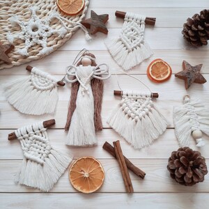 Könnte beinhalten: Eine Sammlung handgefertigter Weihnachtsornamente aus Makramee, darunter Engel, Schneeflocken und geometrische Formen. Die Ornamente sind weiß und beige, mit Holzakzenten und Zimtstangen. Getrocknete Orangenscheiben und Holzsterne ergänzen das festliche Thema.