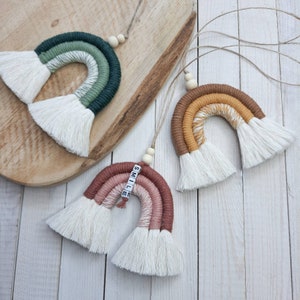 May include: Three colourful macrame rainbows with white tassels. The rainbows are made with different coloured yarn and have wooden beads at the top. The middle rainbow has the word "SMILE" spelled out with white letter tiles.