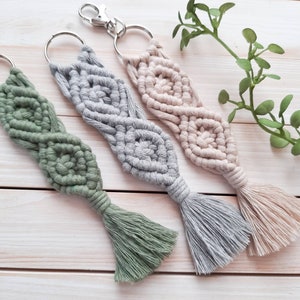 Puede incluir: Tres llaveros de macramé con borlas en verde, gris y beige. Los llaveros están hechos con un patrón de diamante y cuelgan de llaveros de plata.
