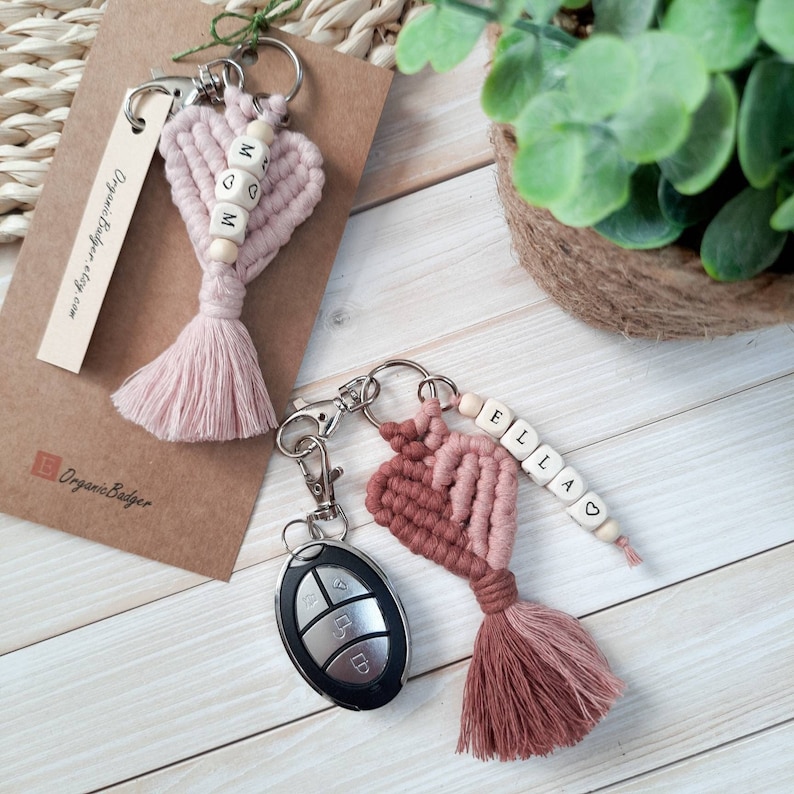 May include: Two pink and red heart-shaped keychains with wooden beads spelling out "MOM" and "ELLA". The keychains have pink and red tassels and are attached to silver key rings.