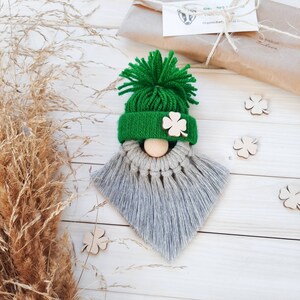 Könnte beinhalten: Ein grüner Zwerg mit einem grauen Bart aus Garn. Der Zwerg trägt eine grüne Strickmütze mit einem Kleeblatt auf der Vorderseite. Der Zwerg sitzt auf einer weißen Holzoberfläche.