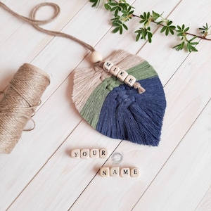 Op de afbeelding: Een blauw, groen en beige macramé blad met een houten kraal en de naam "MIKE" gespeld in houten blokken. De woorden "YOUR NAME" zijn gespeld in houten blokken onder het blad.