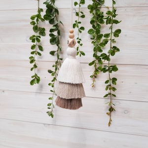 Może przedstawiać: Trójwarstwowa frędzla w kolorze beżowym, jasnobrązowym i białym. Frędzla jest zawieszona na drewnianej koralce z literami "B" i "A".