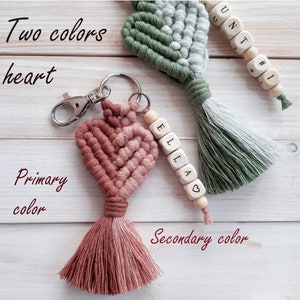 May include: Two-toned heart-shaped keyring with a pink and brown tassel. The keyring is made of woven string and has a silver key ring. The keyring is attached to a wooden bead with the name "Ella" spelled out on it.