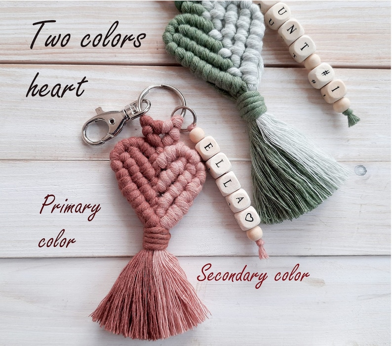 May include: Two-toned heart-shaped keyring with a pink and brown tassel. The keyring is made of woven string and has a silver key ring. The keyring is attached to a wooden bead with the name "Ella" spelled out on it.