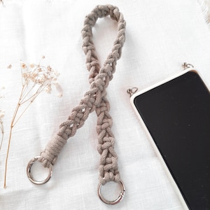 May include: A beige macrame phone strap with silver-colored metal clasps. The woven strap is displayed on a white surface next to a smartphone in a white case. The strap is designed to be attached to a phone case.