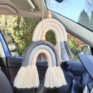 May include: Two macrame rainbows hanging from a rearview mirror in a car. The rainbows are made of beige, blue, and gray yarn and have wooden beads.