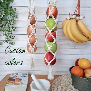 May include: Two white macrame hanging produce bags with onions and apples inside. The bags are hanging on a white wood background. The text "Custom colours" is written in black on the left side of the image. A bowl of oranges and apples is on a woven placemat in the lower right corner of the image.