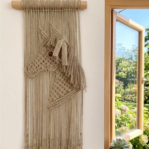 Könnte beinhalten: Ein beigefarbener Makramee-Wandbehang mit einem Pferdekopf-Motiv aus geknüpfter Kordel. Das Stück ist an einem horizontalen Holzstab mit einer Aufhängeschnur befestigt. Die Pferdefigur enthält eine dunkle Perle als Auge und Fransendetails.