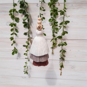 May include: A white, gray, and brown tassel with a wooden bead and a wooden block with the letter "M" on it. The tassel is hanging from a white wall with green vines.