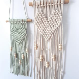 May include: Two macrame wall hangings with heart designs. One is a soft green, the other is off-white. Both feature wooden dowels and beads. The heart shapes are created with knotted rope, and the fringe hangs below.