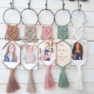 Può includere: Cinque arazzi da parete in macramè con nappe, ognuno con una piccola cornice per foto con un'immagine diversa. Una cornice ha una carta con il testo "Mia Will You be my Bridesmaid?"