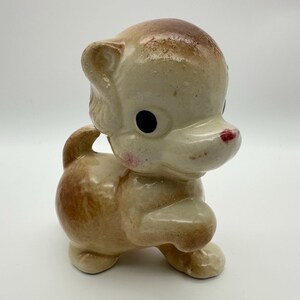 May include: A vintage ceramic figurine of a bear cub in a playful pose. The bear has a cream-colored body with brown accents on its head, back, and paws. It features black eyes, a red nose, and pink cheeks. The figurine is a collectible item.