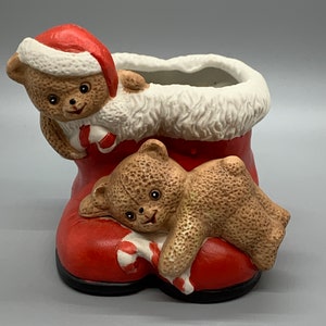 May include: A ceramic Christmas boot planter with two teddy bears inside. The boot is red with white fur trim and a candy cane. The bears are brown with black eyes and noses.