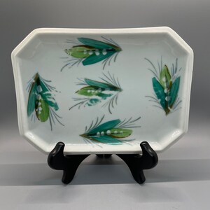 May include: A white ceramic rectangular dish with a decorative design of green and brown feathers. The dish is on a black stand.