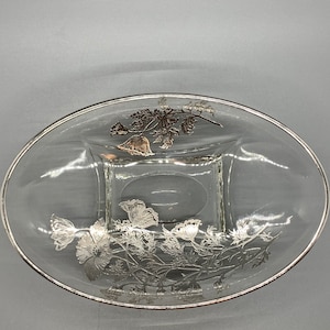 May include: A clear glass oval dish with a silver floral design. The design features leaves and flowers.
