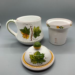 May include: White ceramic teacup and lid with a green and yellow vine design. The lid has a small green knob. The teacup has a handle.