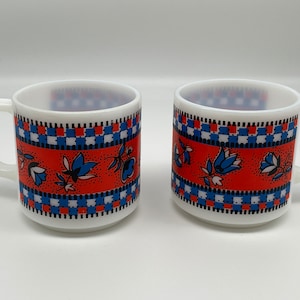 May include: Two white ceramic mugs with a red, blue, and white floral pattern. The mugs have a checkered pattern around the bottom.
