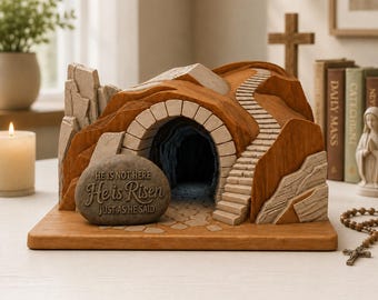 May include: A decorative tomb sculpture featuring a brown rocky mountain with a stone staircase and a dark cave entrance. A gray stone in the foreground is inscribed with the text: HE IS NOT HERE, He is Risen, JUST AS HE SAID.