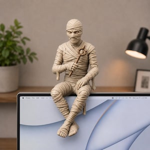 May include: A beige mummy figurine wrapped in bandages, sitting with legs crossed. The figure holds a small, brown, circular-topped staff in one hand. The mummy has visible facial features, including eyes and a nose, and is perched on the top edge of a monitor.