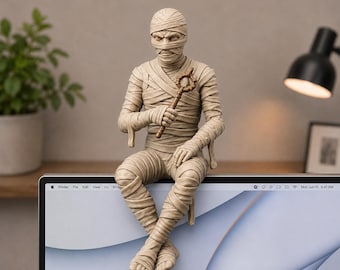 May include: A beige mummy figurine wrapped in bandages, sitting with legs crossed. The figure holds a small, brown, circular-topped staff in one hand. The mummy has visible facial features, including eyes and a nose, and is perched on the top edge of a monitor.