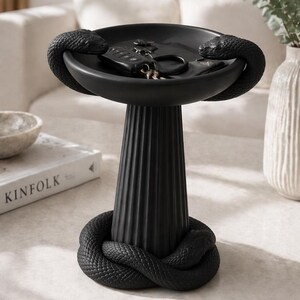 May include: A black decorative tray with a pedestal base, featuring a coiled snake design at the base and two snakes encircling the bowl. The bowl holds keys and a key fob. The tray is made of a matte black material.